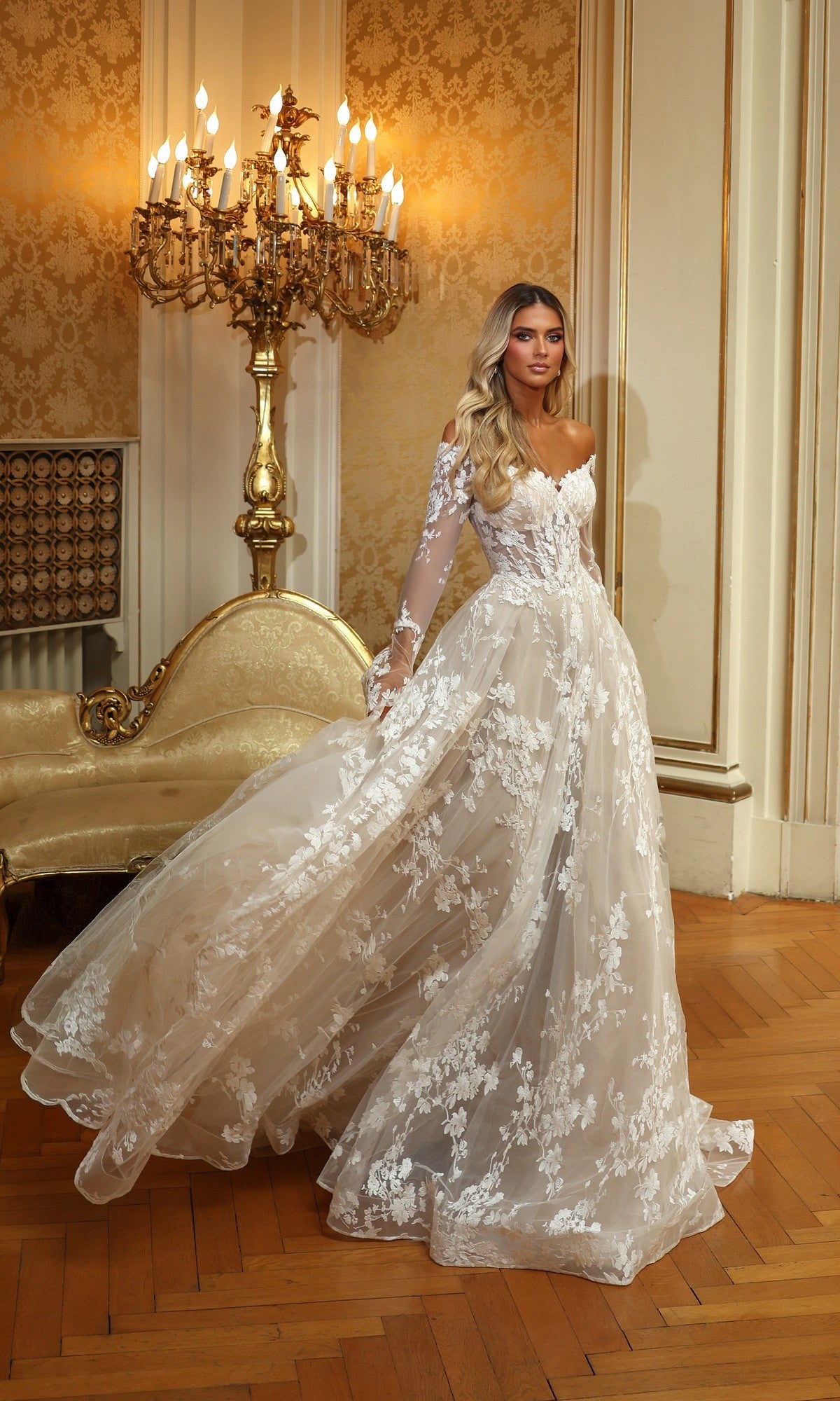 A woman wears the Alyce 7150 long ivory solid dress with lace, sheer sleeves, and ruffle details, standing in an ornate room with gold accents and a vintage chandelier.